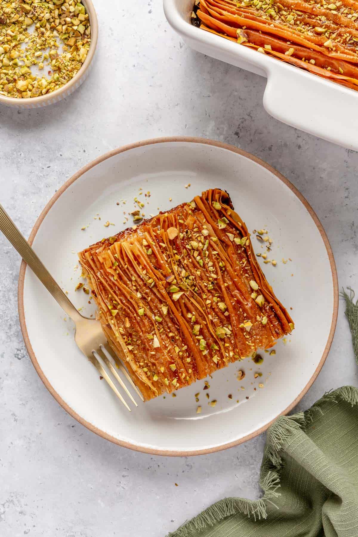 Serving of crinkle cake on a plate showing it's golden brown crispy top with crushed pistachios on top.