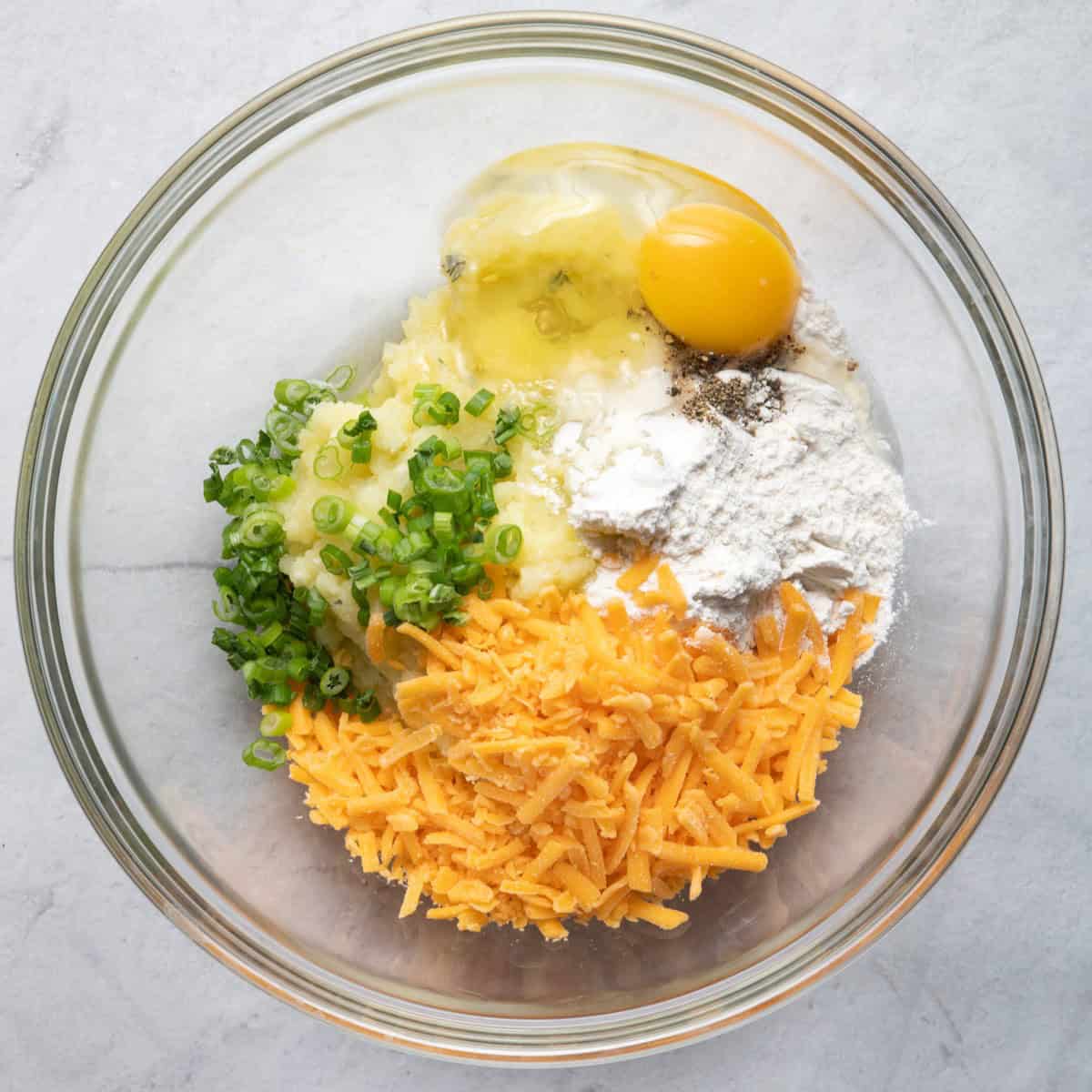 Mashed potatoes in a bowl with egg, cheese, flour, green onions, salt and pepper.