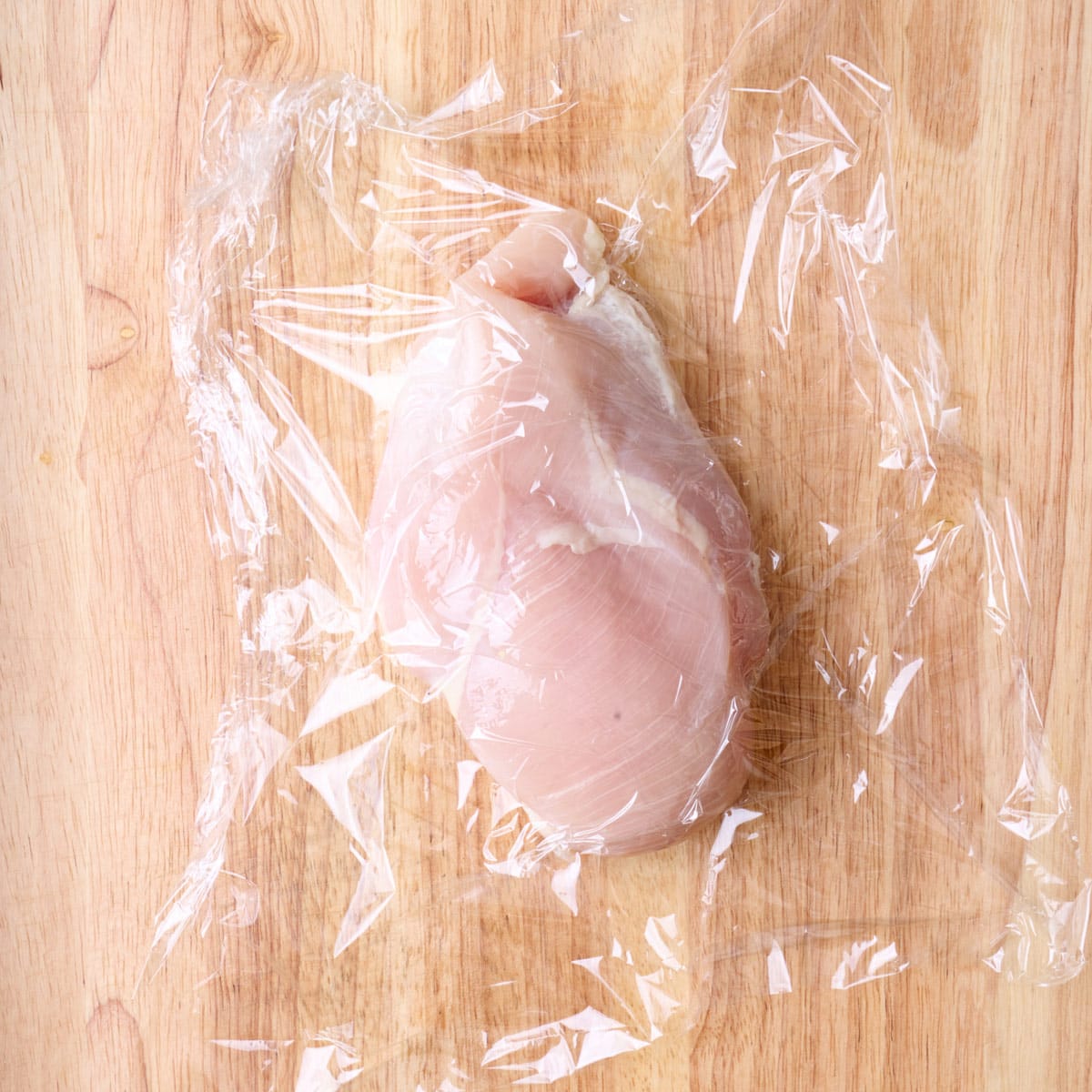 Chicken breast on a cutting board cover with plastic wrap.