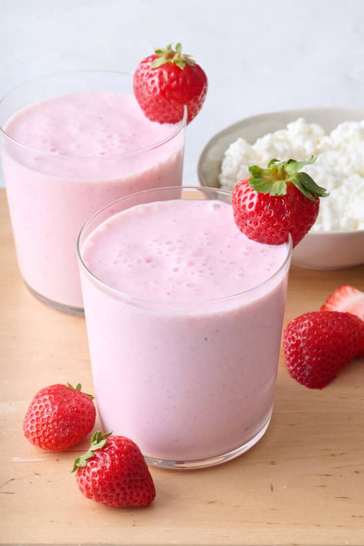 Two glasses filled with strawberry cottage cheese smoothie with whole strawberry on rim of glasses and bowl of cottage cheese nearby.