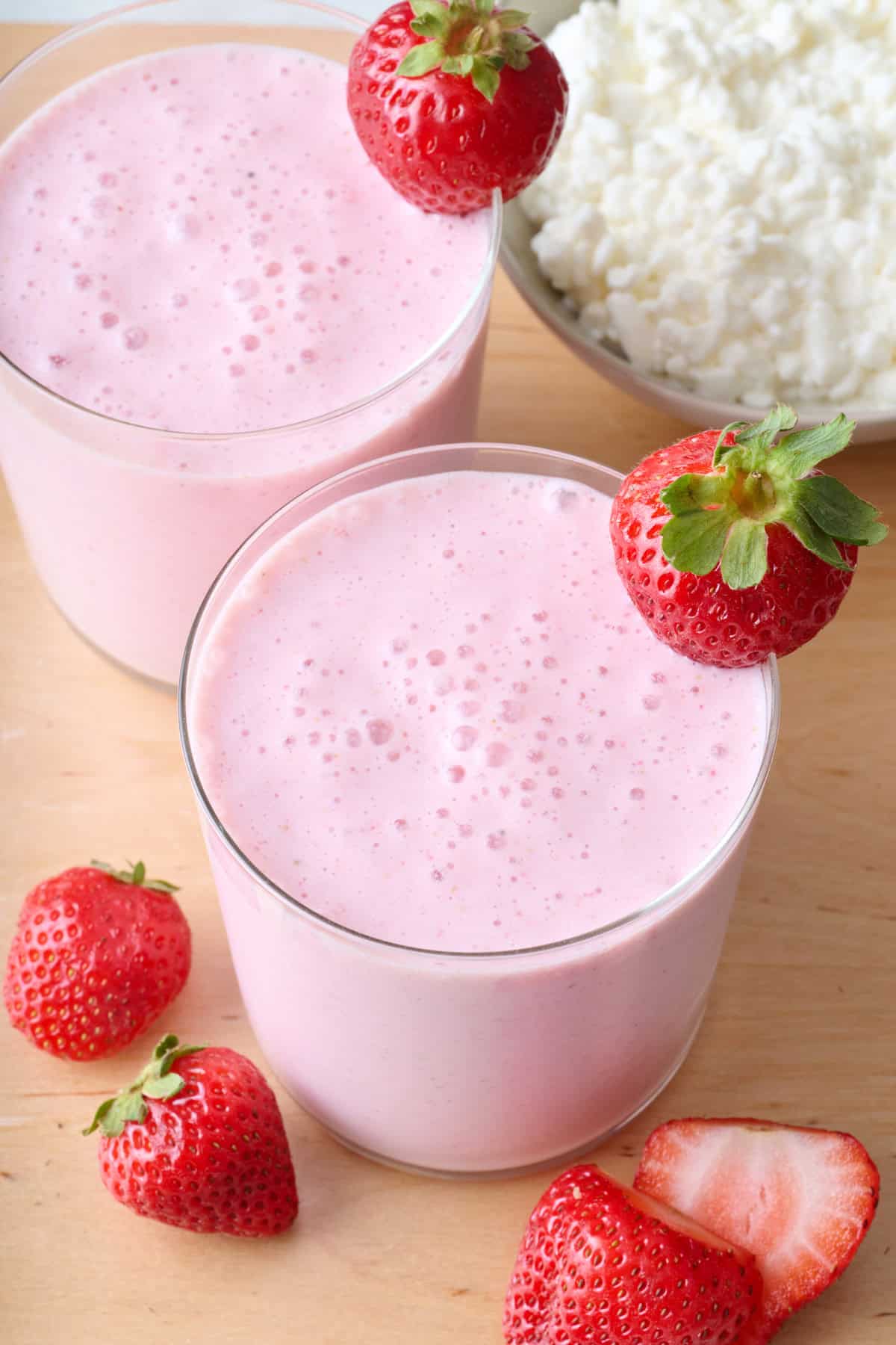 Two glasses filled with cottage cheese smoothie with whole strawberry on rim of glasses and bowl of cottage cheese nearby.