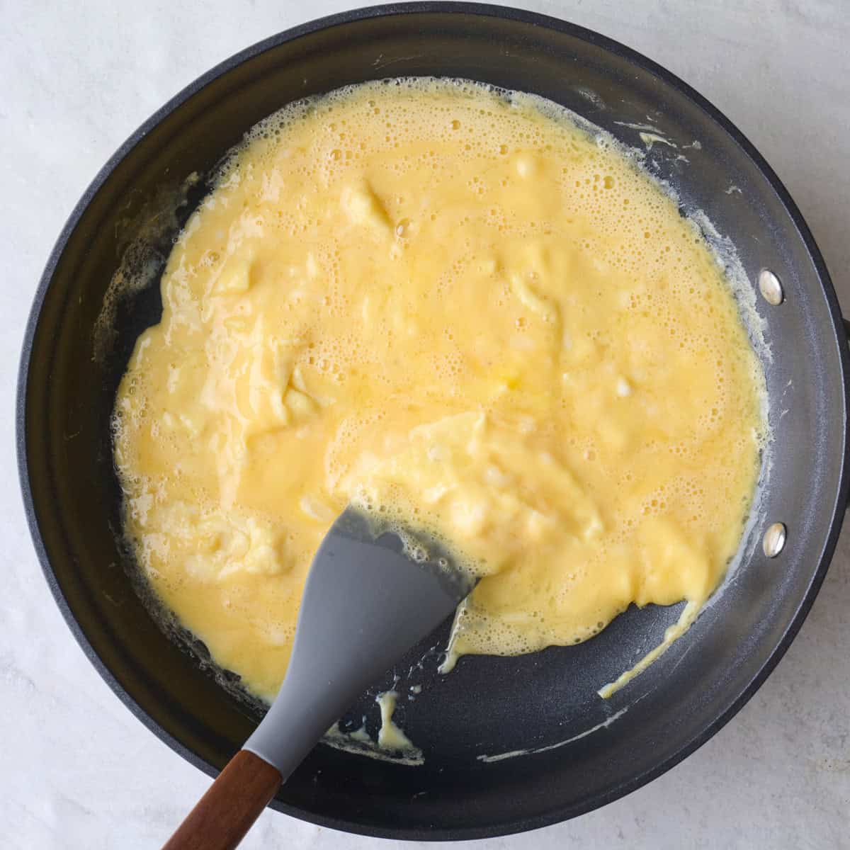 Spatula pushing partially cooked beaten eggs in a pan towards the center.