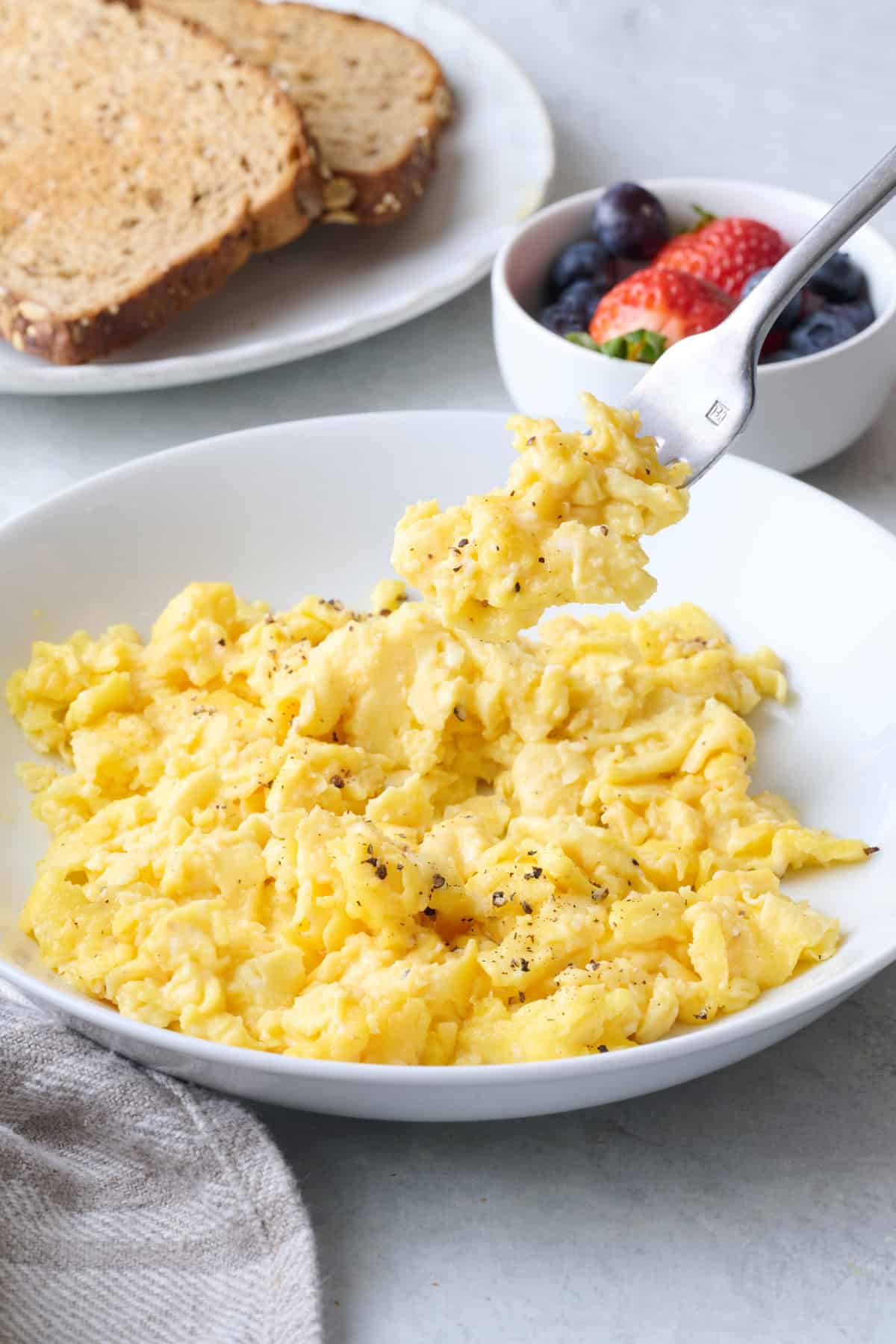 Fork lifting up a bite of scrambled eggs with a plate of toast and dish of fresh berries nearby.