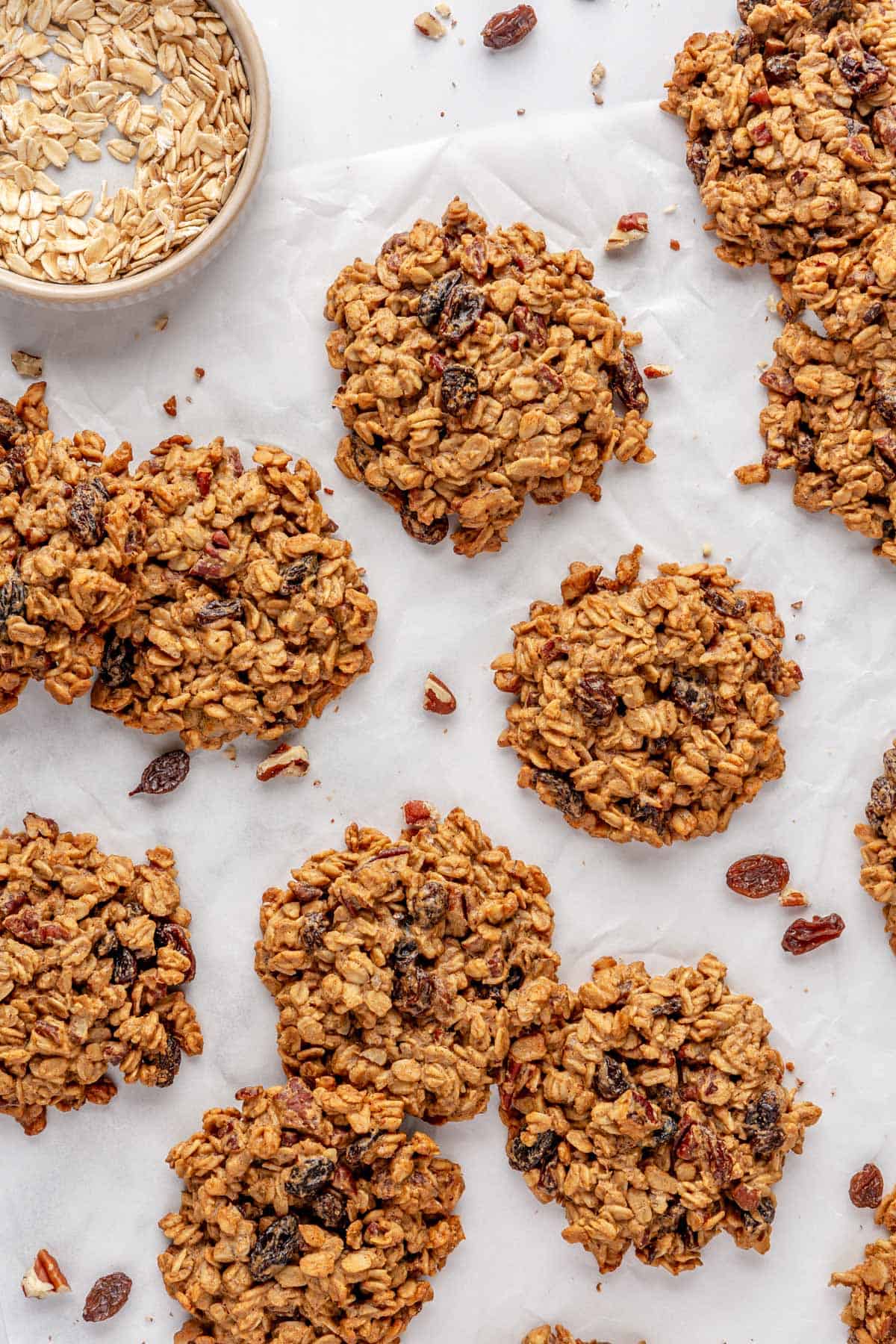 Cookies on parchment paper with a small dish of oats nearby and just a few pecans and raisins around.