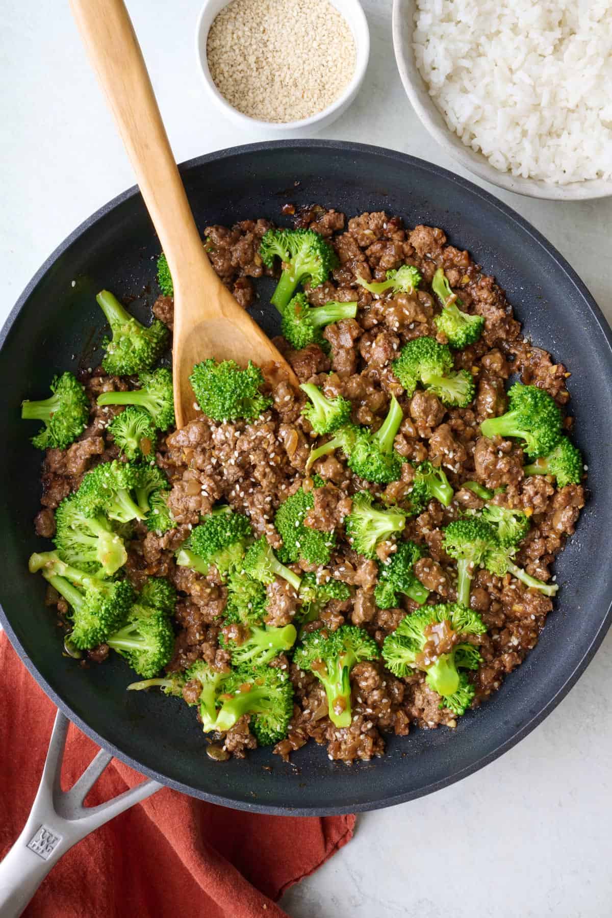 Ground beef and broccoli in the skillet garnished with sesame seeds, wooden spoon dipped in.