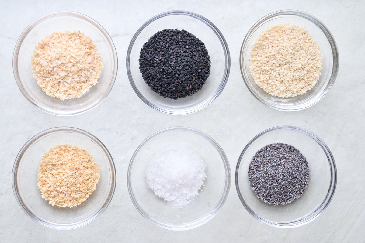 Ingredients for recipe in individual bowls: white sesame seeds, black sesame seeds, dried minced garlic, dried minced onion, poppy seeds, and sea salt.