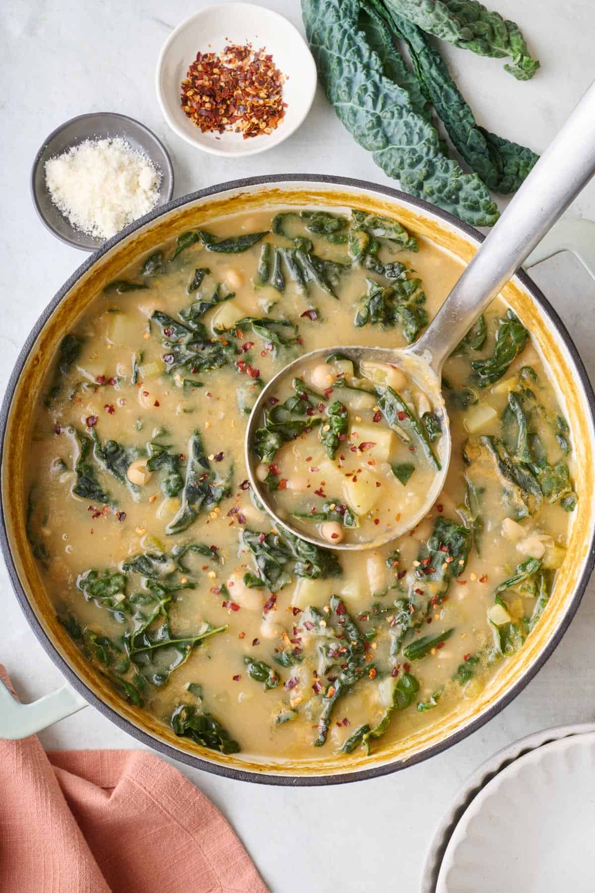 Kale Soup in pot with ladle dipped inside, small dish of red pepper flakes and parmesan cheese nearby.