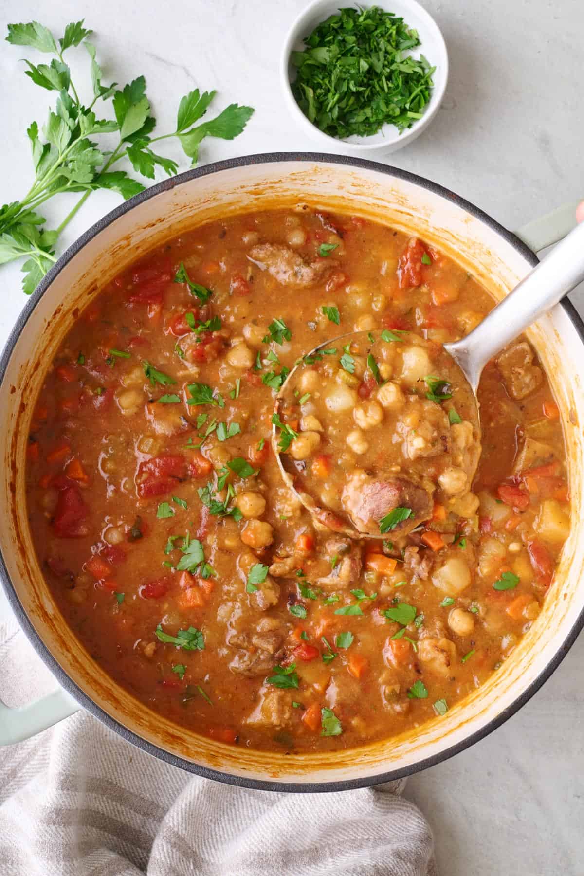 Lamb Stew in pot garnished with parsley and with ladle dipped inside, fresh herbs nearby.