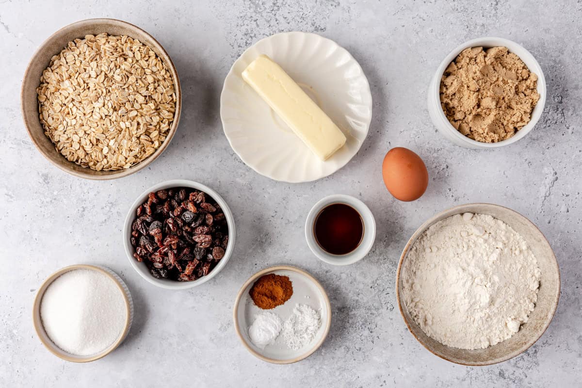 Ingredients for recipe in individual dishes: oats, raisins, sugar, butter, vanilla extract, cinnamon, baking powder, baking soda, flour, egg, and brown sugar.