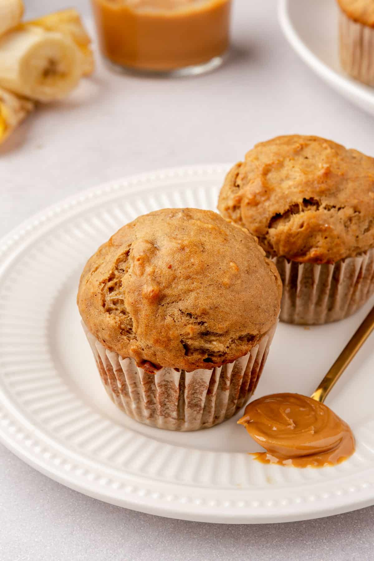 Two peanut butter banana muffins on a plate with a small spoon of peanut butter resting nearby.