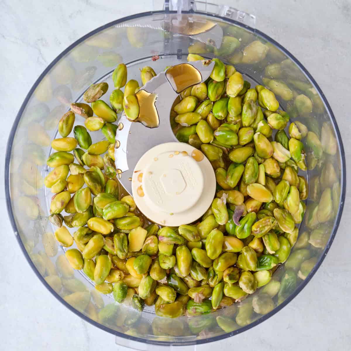 Peeled pistachios in the bowl of a food processor with maple syrup and salt.