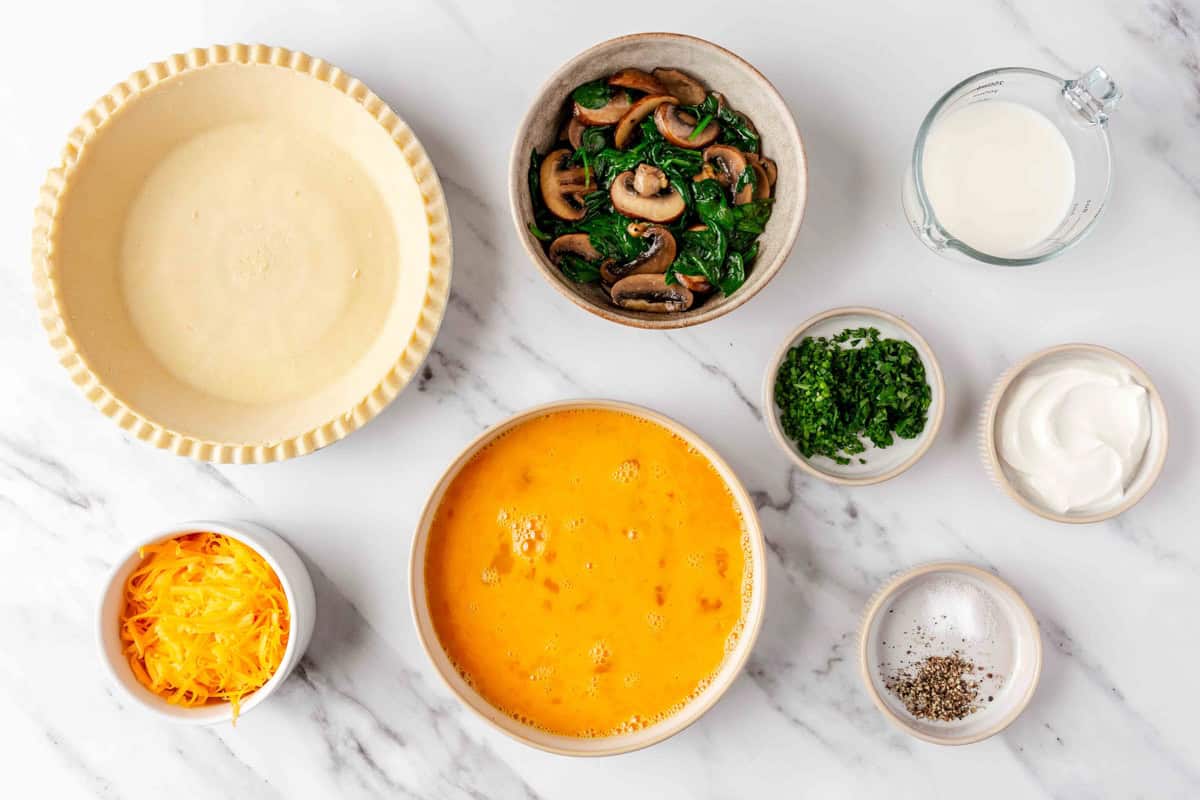 Ingredients for recipe in individual dishes: pie crust, shredded cheese, spinach mushroom mixture, scrambled eggs, chopped chives, milk, yogurt and seasoning.