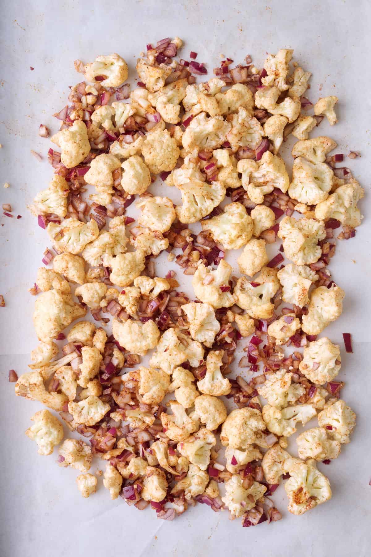 Cauliflower florets and diced onion coated in spices and oil on a baking sheet before roasting.