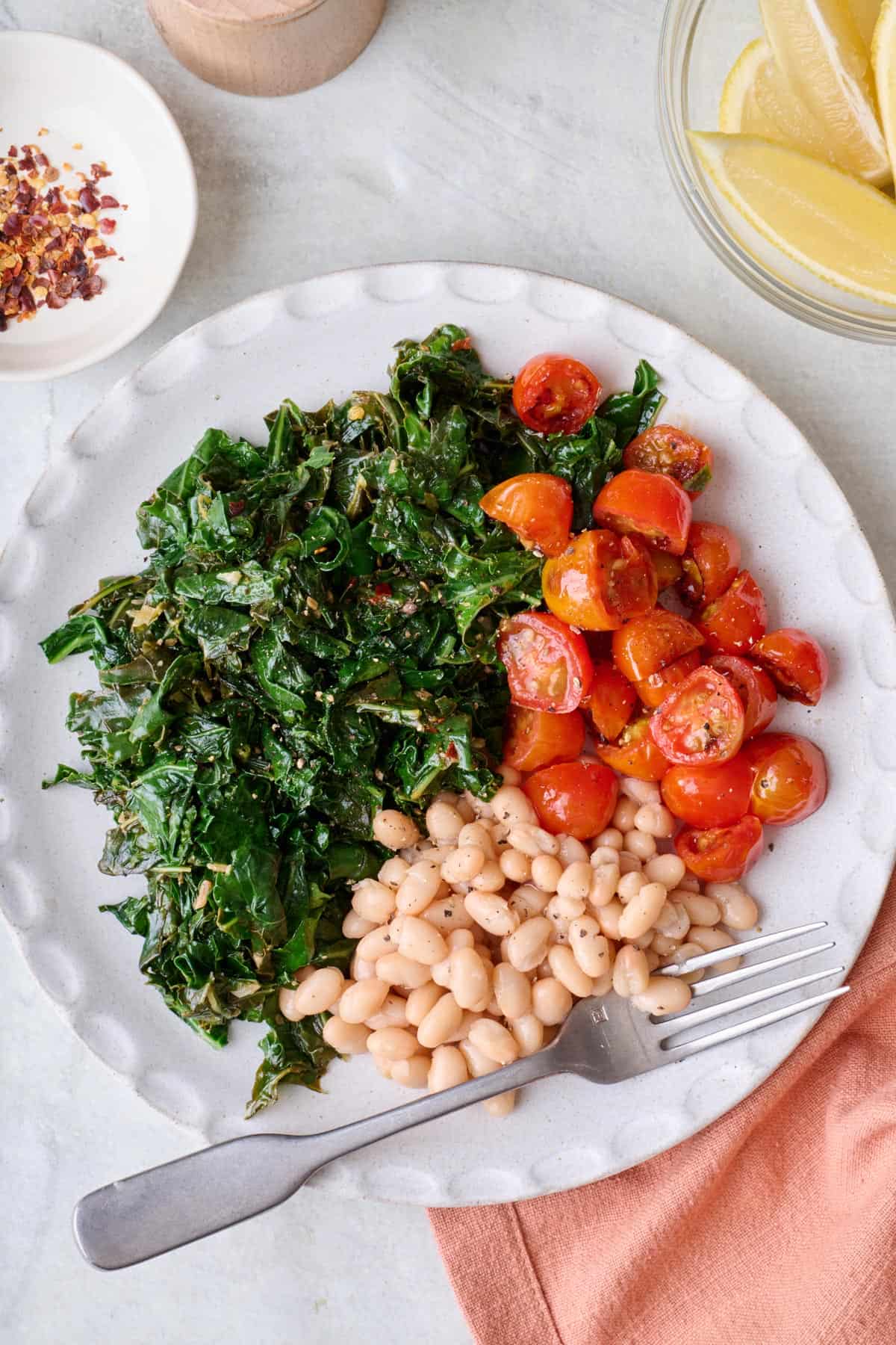 Serving of kale in a bowl with small side of halved sauteed cherry tomatoes and small portion of white beans.