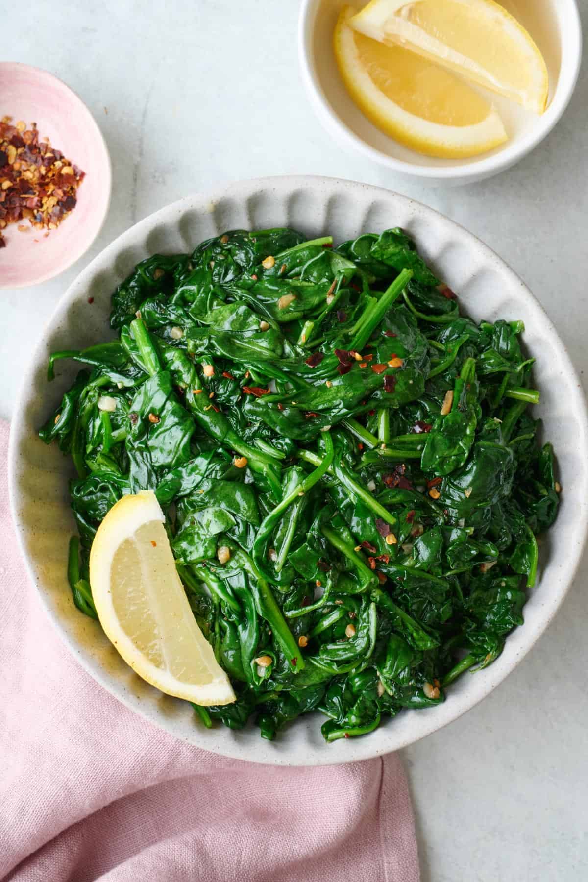 Sauteed spinach in a shallow serving bowl with lemon wedges and small dish of extra lemons nearby.