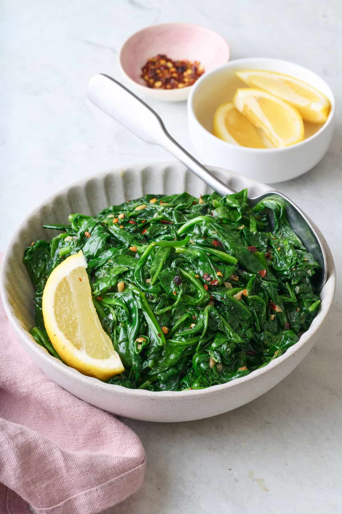 Sauteed spinach in a shallow serving bowl with lemon wedges and small dish of extra lemons nearby.