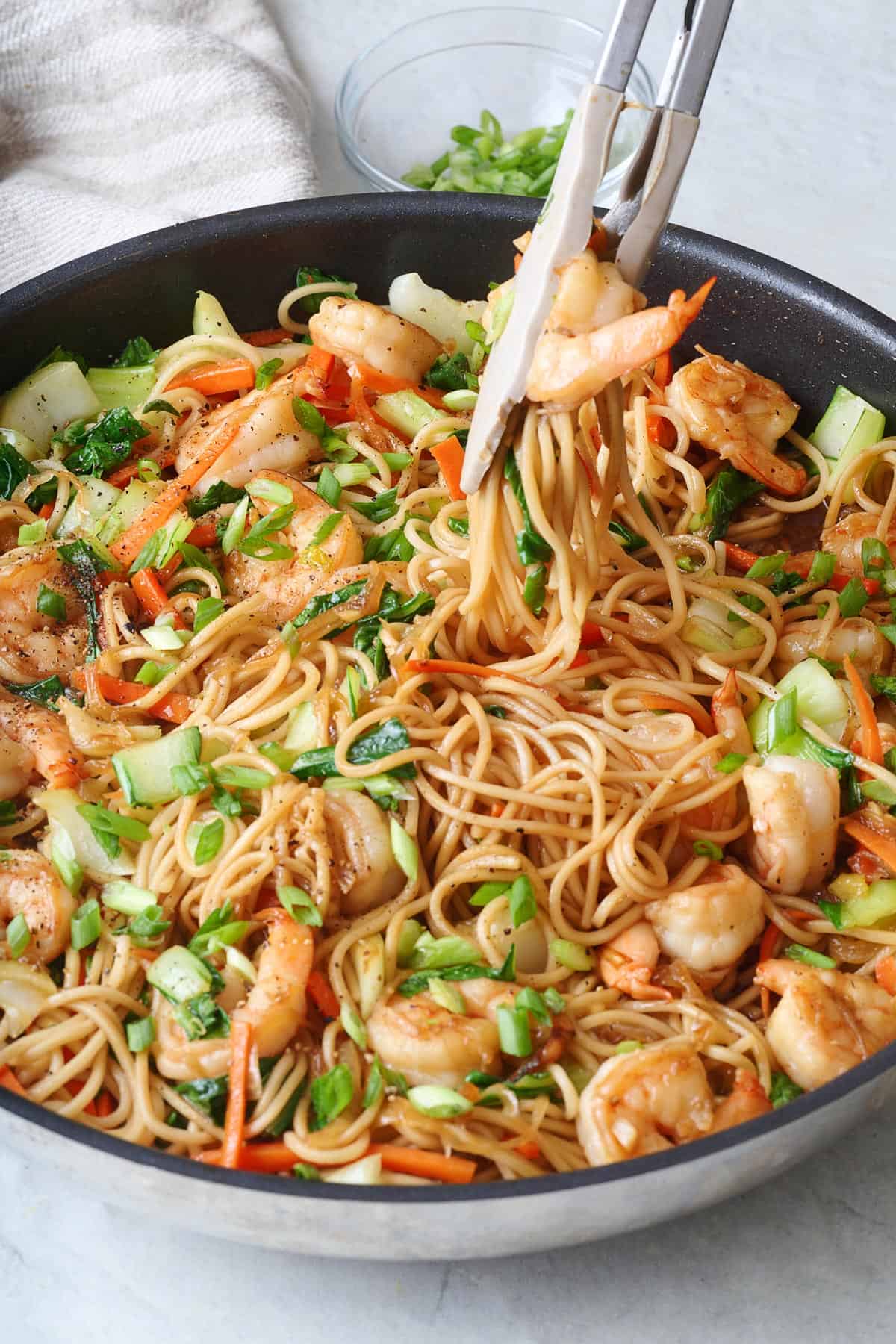 Tongs lifting up shrimp lo mein from a skillet.