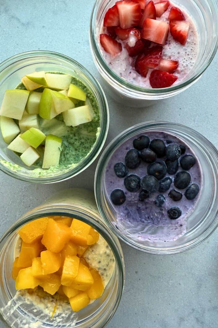 Blended overnight oats in 4 jars with 4 different fresh fruit flavor varieties: apple and spinach, strawberry, mango, and blueberry.