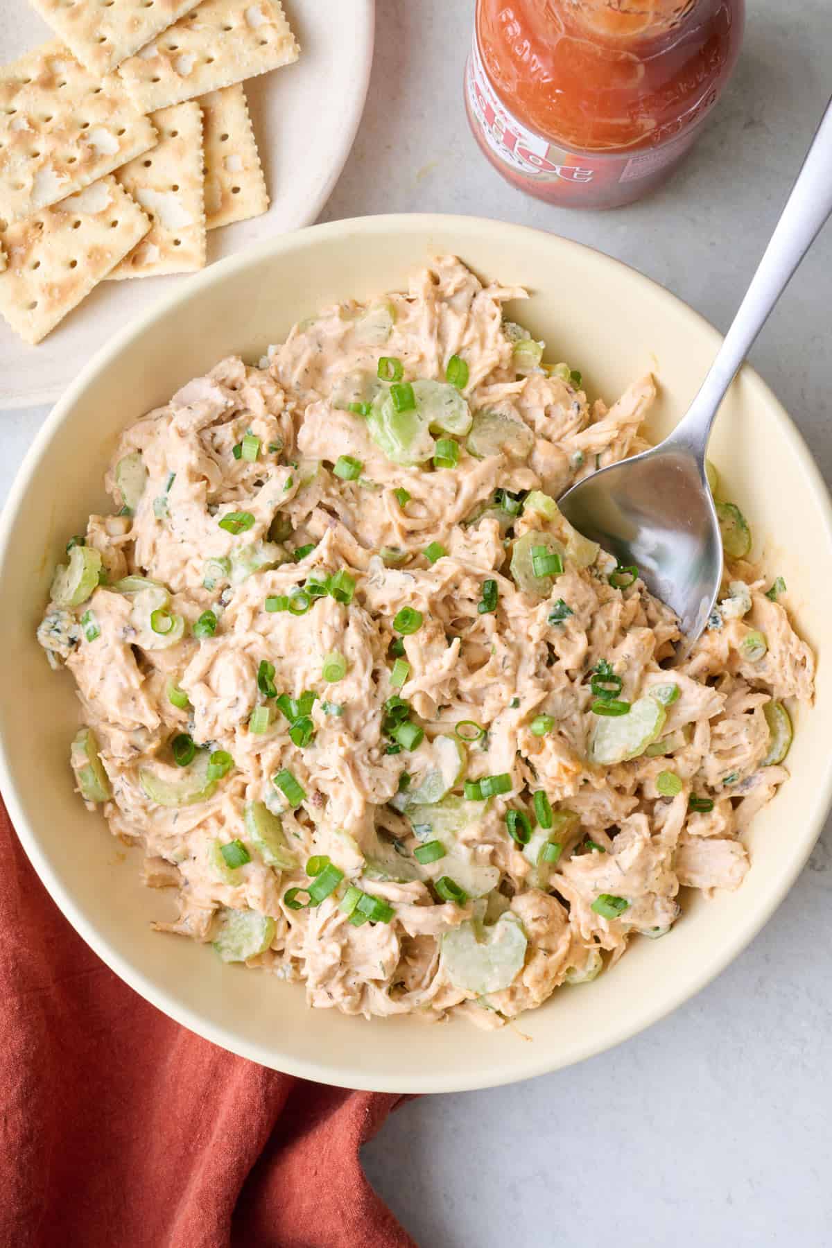 Buffalo chicken salad in a serving bowl garnished with extra green onions with serving spoon dipped inside, a few crackers and lettuce cups nearby.