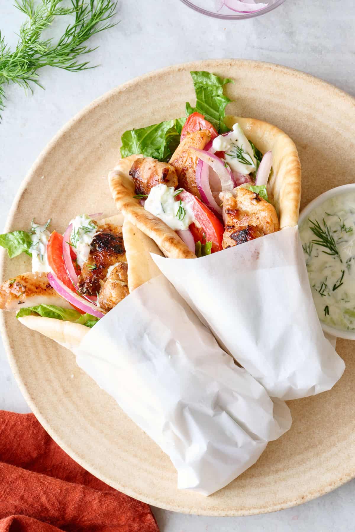 Two assembled gyros folded on a plate and wrapped with paper with small dish of tzatziki sauce nearby.