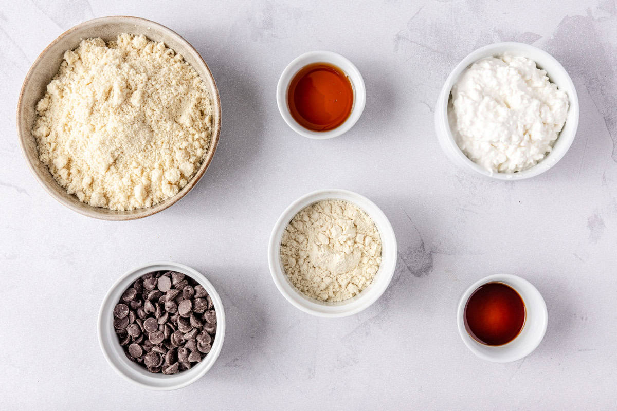 Ingredients to make recipe in individual bowls: almond flour, chocolate chips, vanilla, maple syrup, protein powder, and cottage cheese.