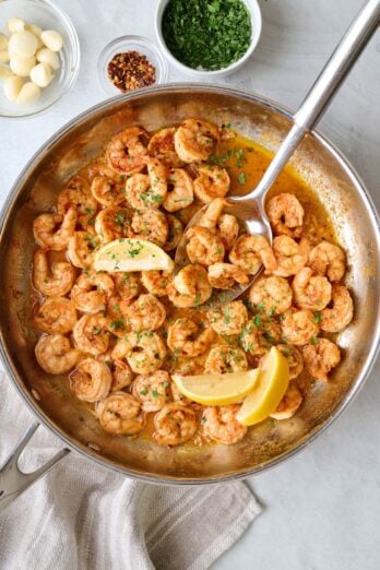 Shrimp in pan garnished with lemon wedges and chopped fresh parsley, whole garlic cloves or bulb nearby, large spoon dipped inside.