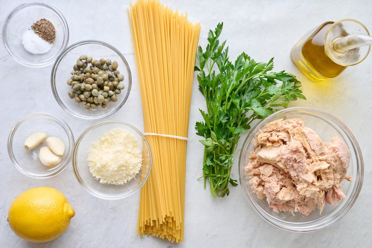 Ingredients for recipe: salt and pepper, garlic, capers, parmesan, lemon, spaghetti noodles, fresh parsley, canned tuna in a bowl, and oil.