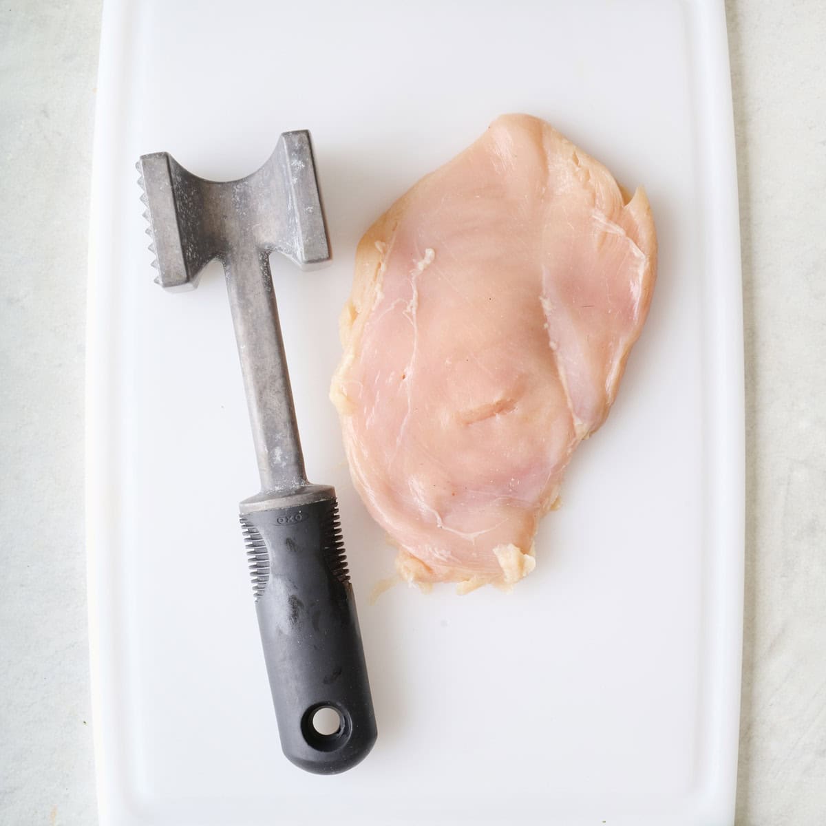 One chicken breast on a white cutting board with meat mallet after pounding it to ½ inch thickness.