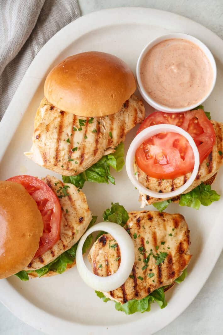 Four grilled chicken sandwiches on a platter with toppings and a side of burger sauce.