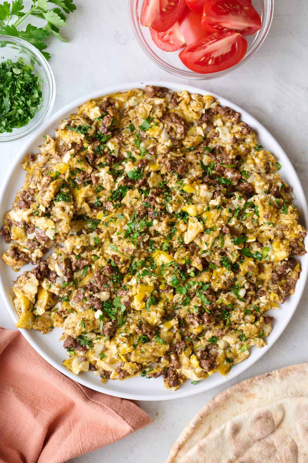 Ground beef and eggs on a plate garnished with fresh parsley with a small dish of quartered tomatoes nearby.