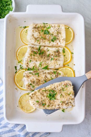 Spatula lifting up a baked cod fillet from baking dish.