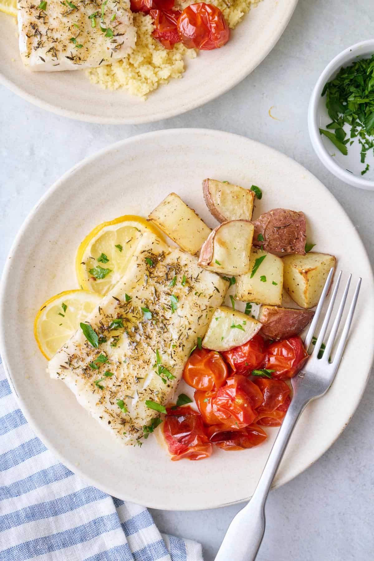 Baked cod on a plate with roasted potatoes and roasted cherry tomatoes.