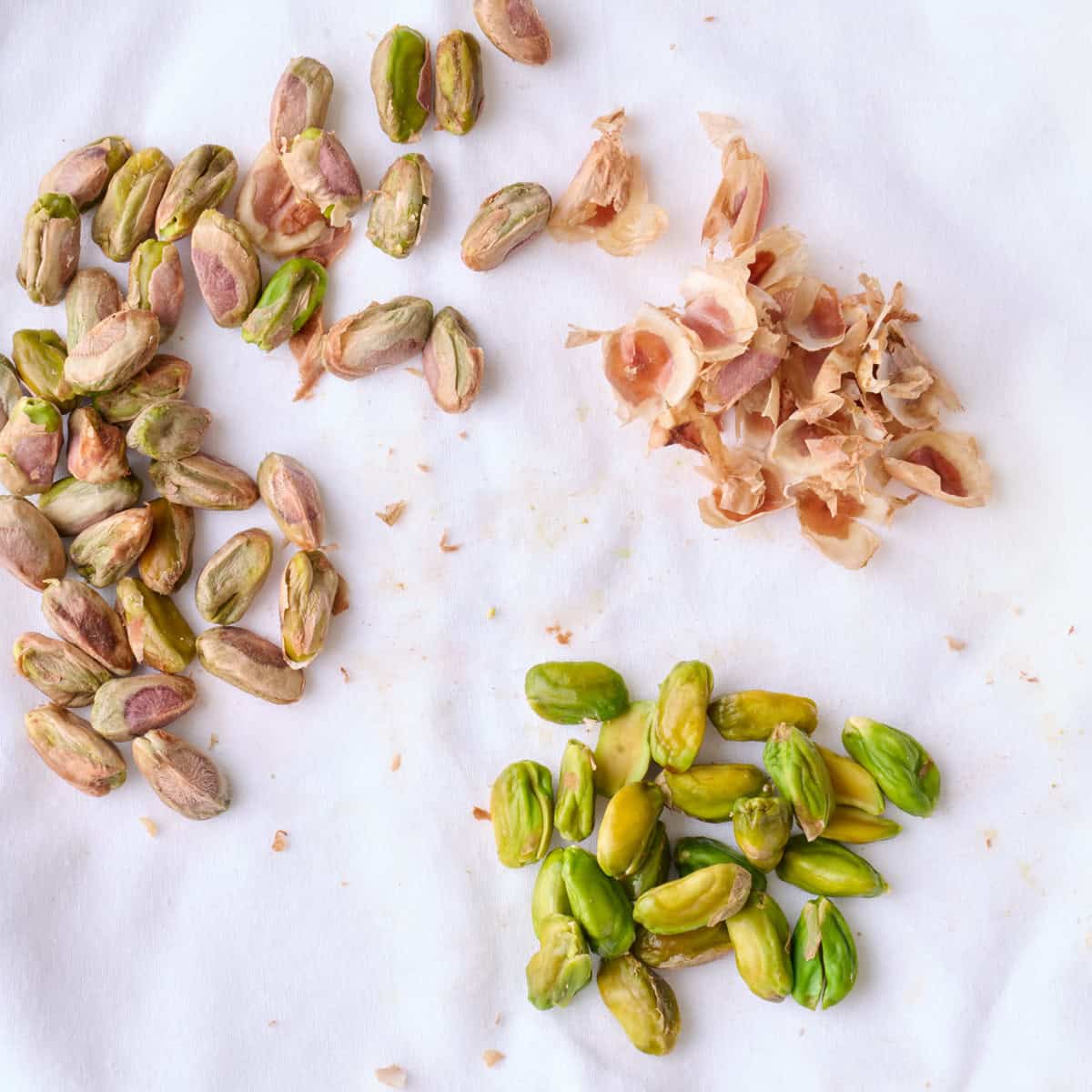 Pistachios on a white cloth getting peeled, some of the peeled already with their skin on the side and others unpeeled.