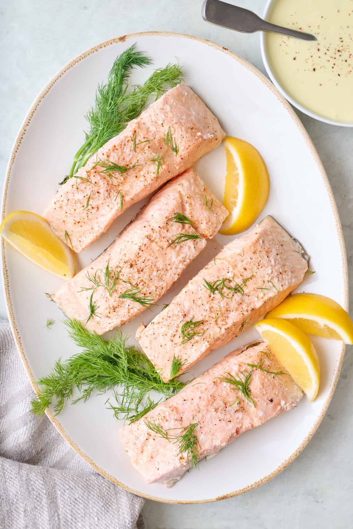 Platter with four poached salmon fillets garnished with lemon wedges, fresh dill and a side of hollandaise sauce.