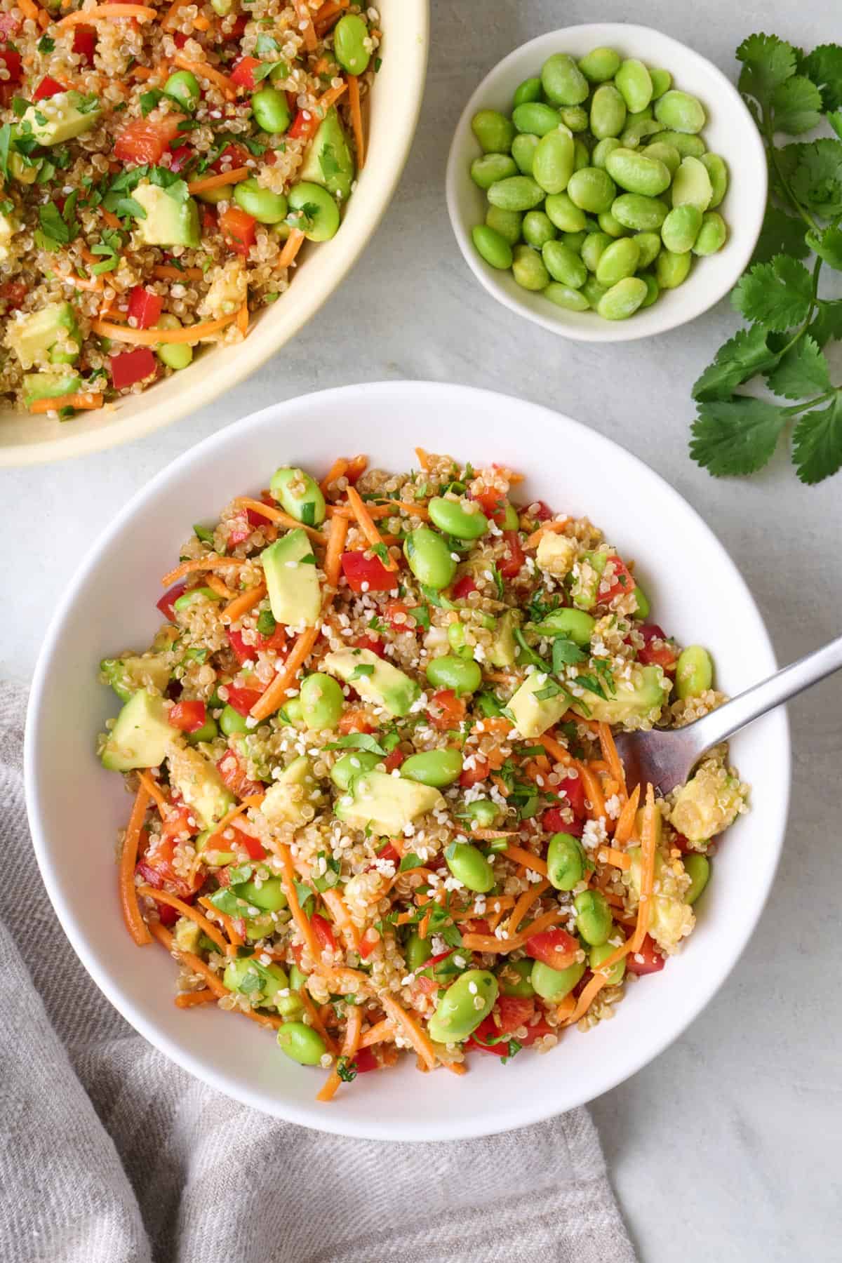 Serving of quinoa salad with edamame and avocado in a bowl with serving dish of more nearby.