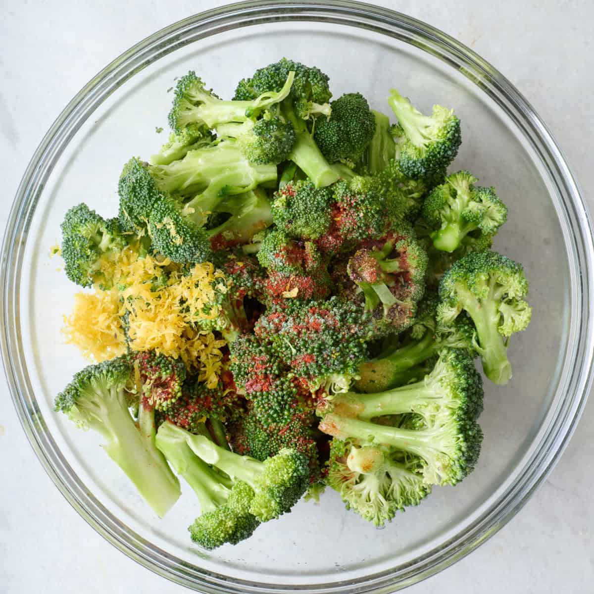 Broccoli florets in a bowl with spices, oil, lemon zest and juice.