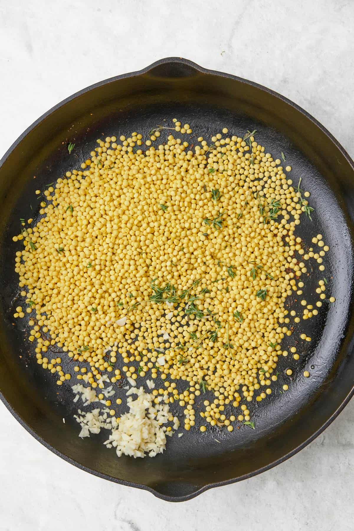 Pearled couscous, herbs, and garlic in a skillet.