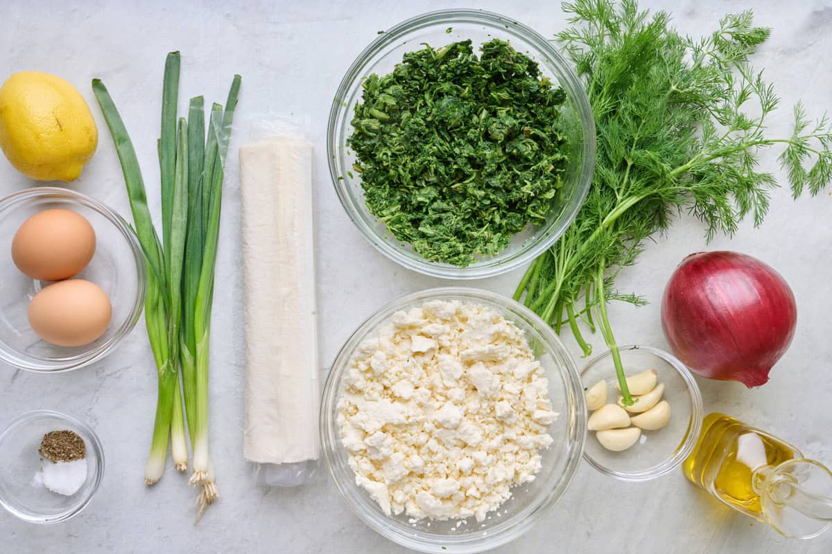 Ingredients for recipe: lemon, eggs, salt and pepper, green onions, phyllo dough, frozen spinach, crumbled feta, fresh dill, garlic, red onion, and olive oil.