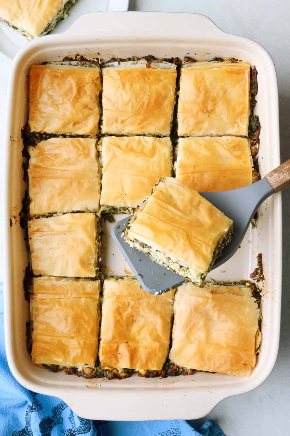 Spanakopita in baking dish with a couple slices removed and spatula lifting one piece.