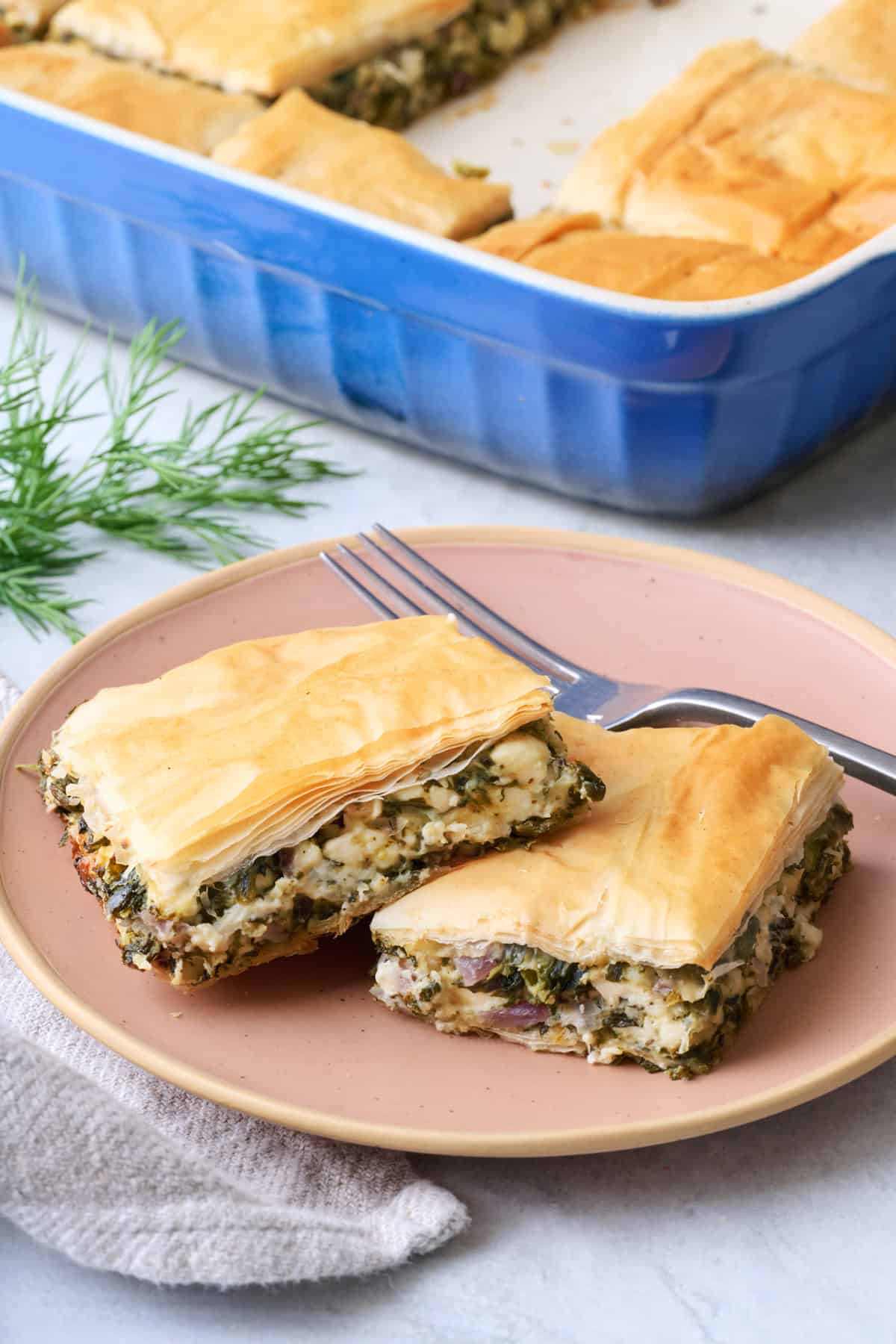 Two pieces of spanakopita on a plate with baking dish nearby.