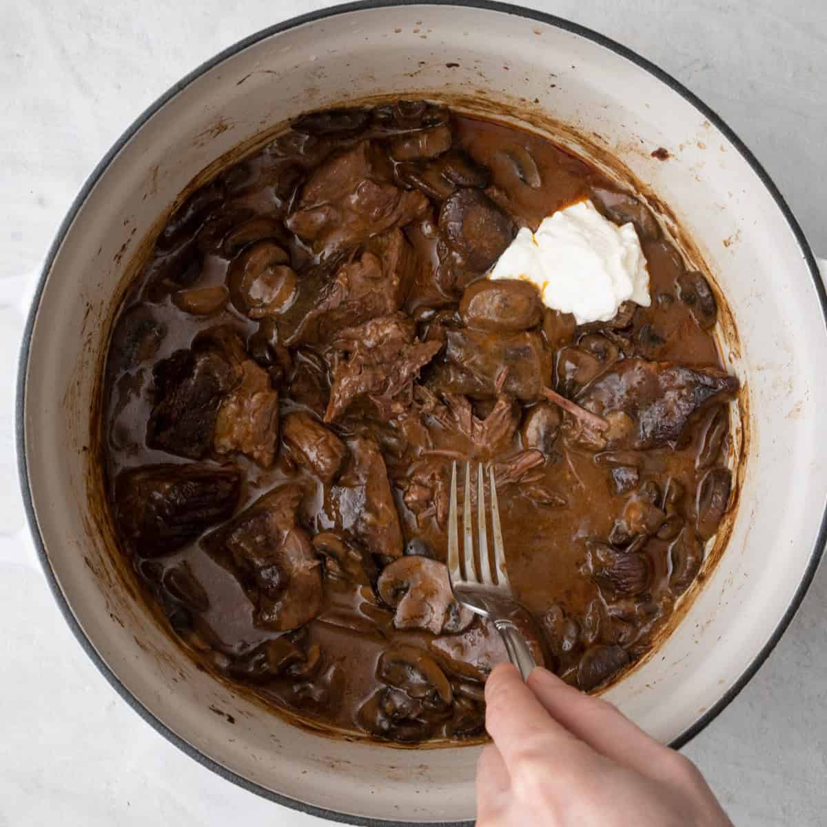 Stroganoff after combined and cooked to show tender beef with fork mashing and Greek yogurt added on top before stirred in.