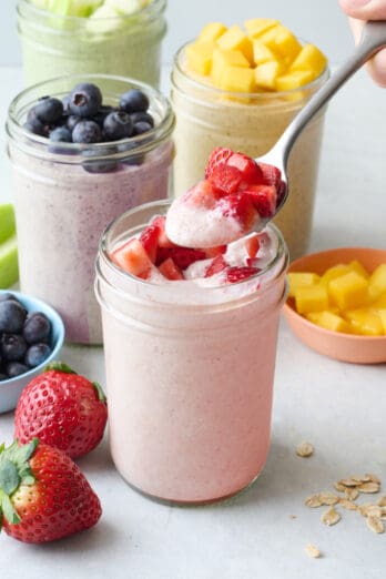 Strawberry blended overnight oats in a small jar topped with fresh strawberries with a spoon lifting some up. More blended overnight oat flavors nearby.