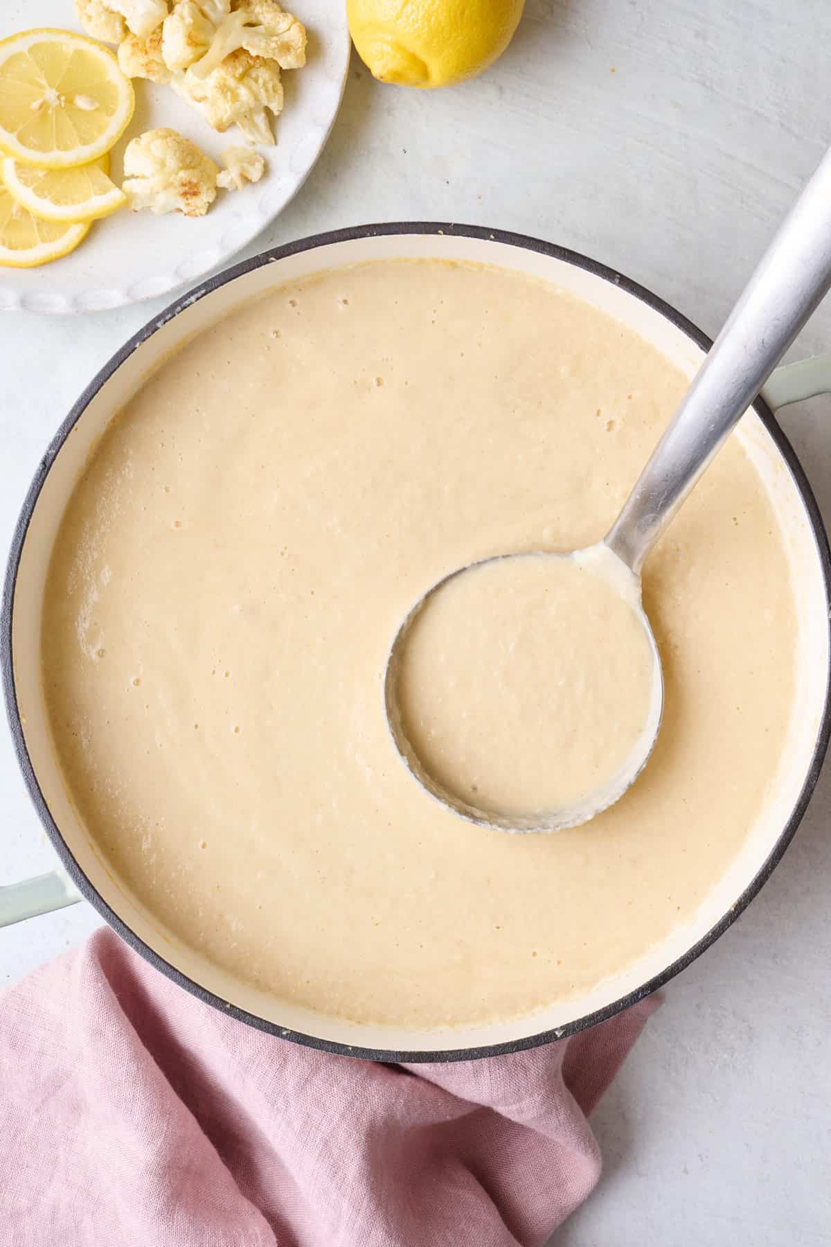 Creamy Cauliflower Soup in pot with ladle dipped inside, small dish of extra roasted cauliflower pieces and lemon nearby