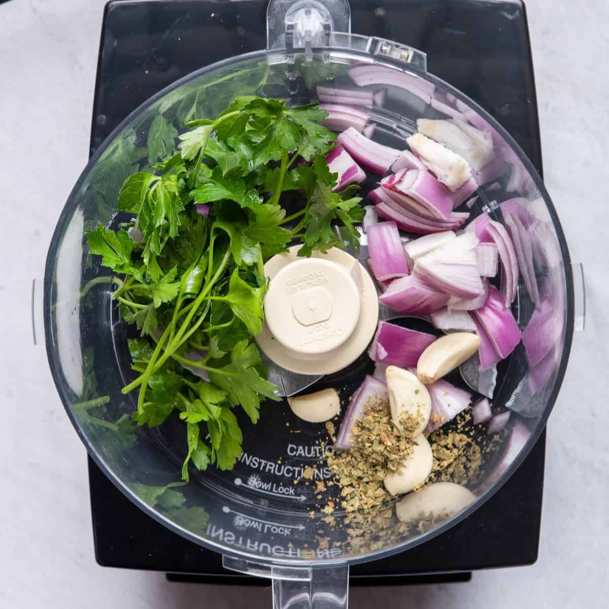 Onions, garlic, herbs, and fresh parsley in the bowl of a food processor.