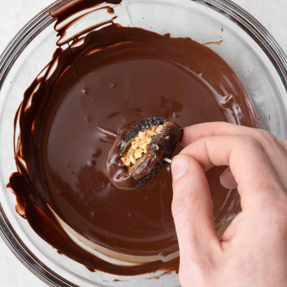 Hand holding peanut butter and nut stuffed date by toothpick and dipping into chocolate.