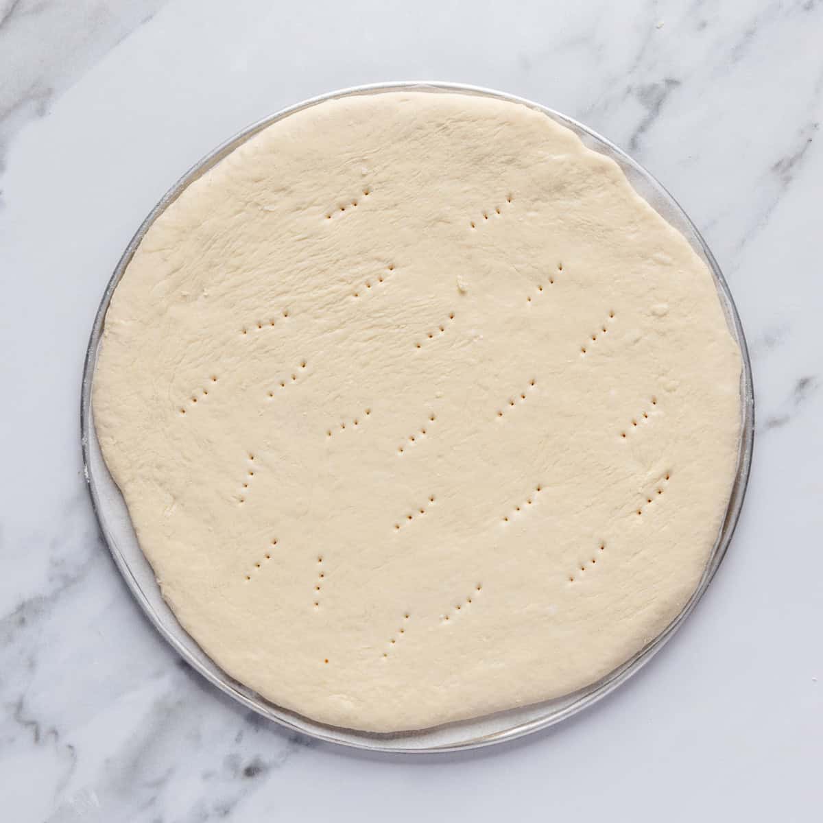 Rolled out dough on a round pizza pan after poking with a fork.