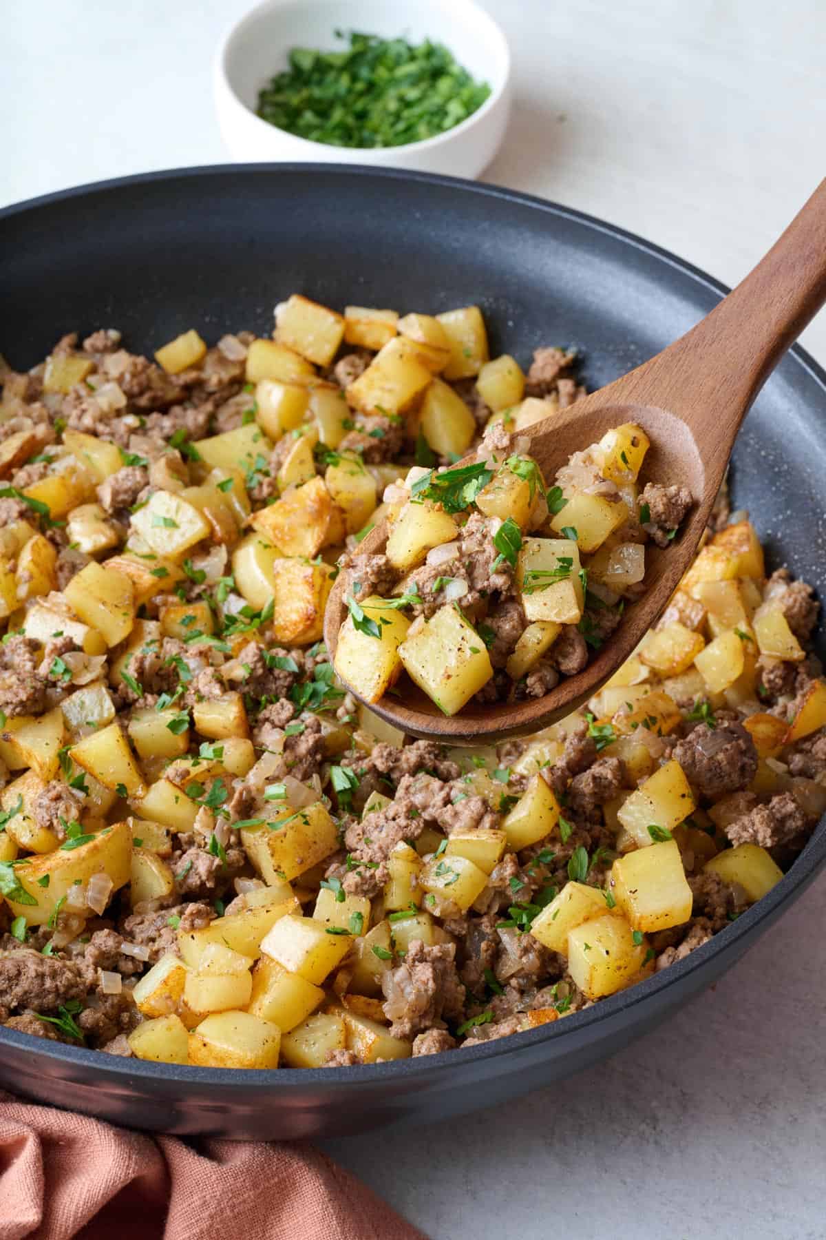 Ground beef and potatoes in the skillet garnished with parsley and a serving spoon lifting up a spoonful.