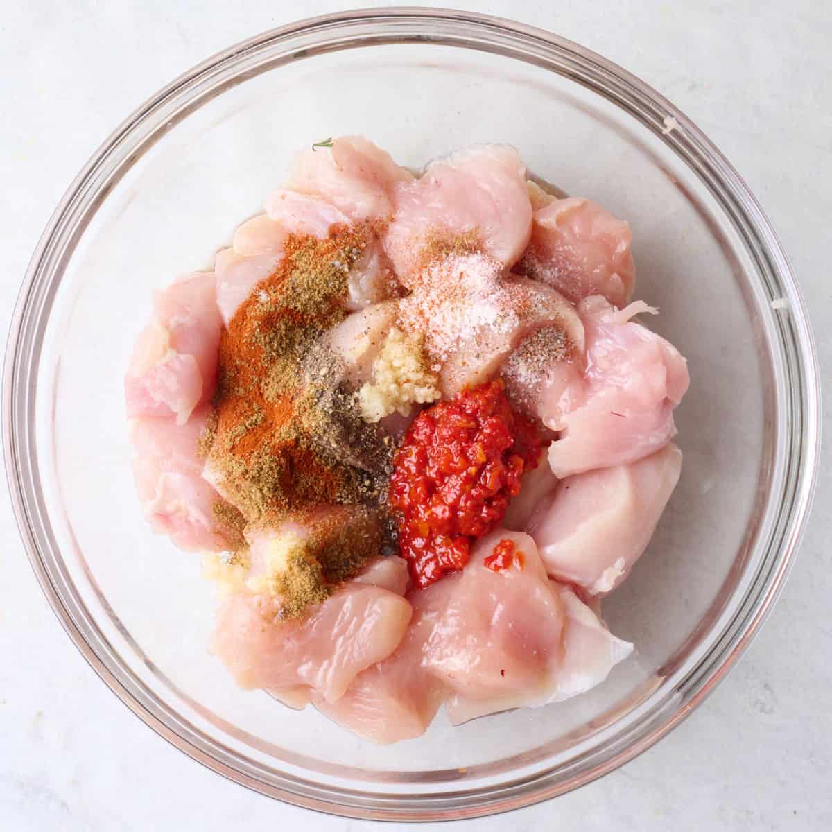 Chicken breast cubes in a bowl with spices, garlic, and harissa.