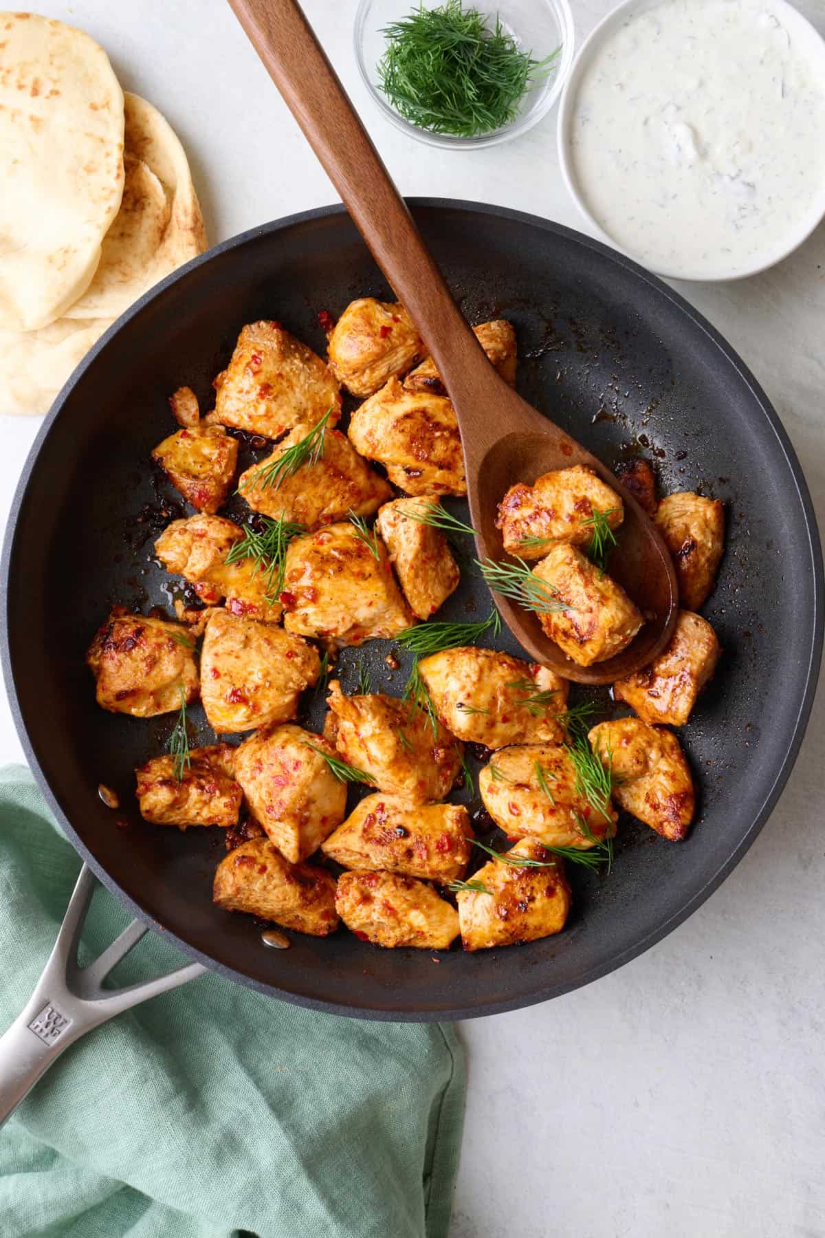 Harissa chicken in skillet garnished with fresh dill, naan and yogurt sauce nearby, serving spoon dipped inside.