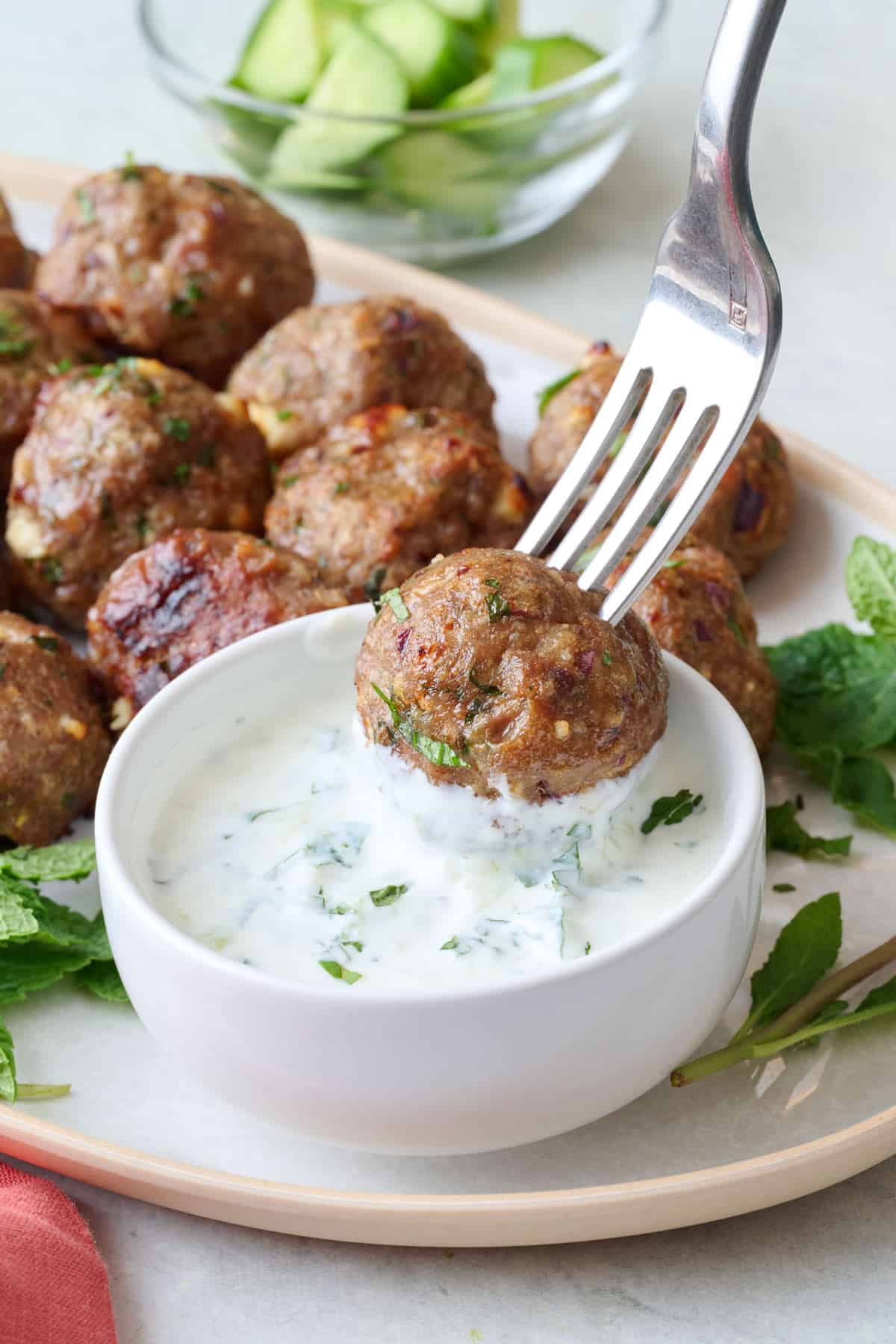 Fork dipping a lamb meatball into sauce on a platter with more meatballs.